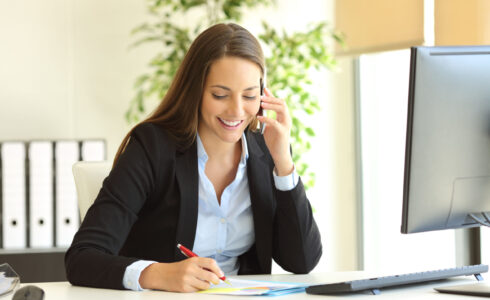 Happy businesswoman calling on mobile phone and taking notes on a desk at office